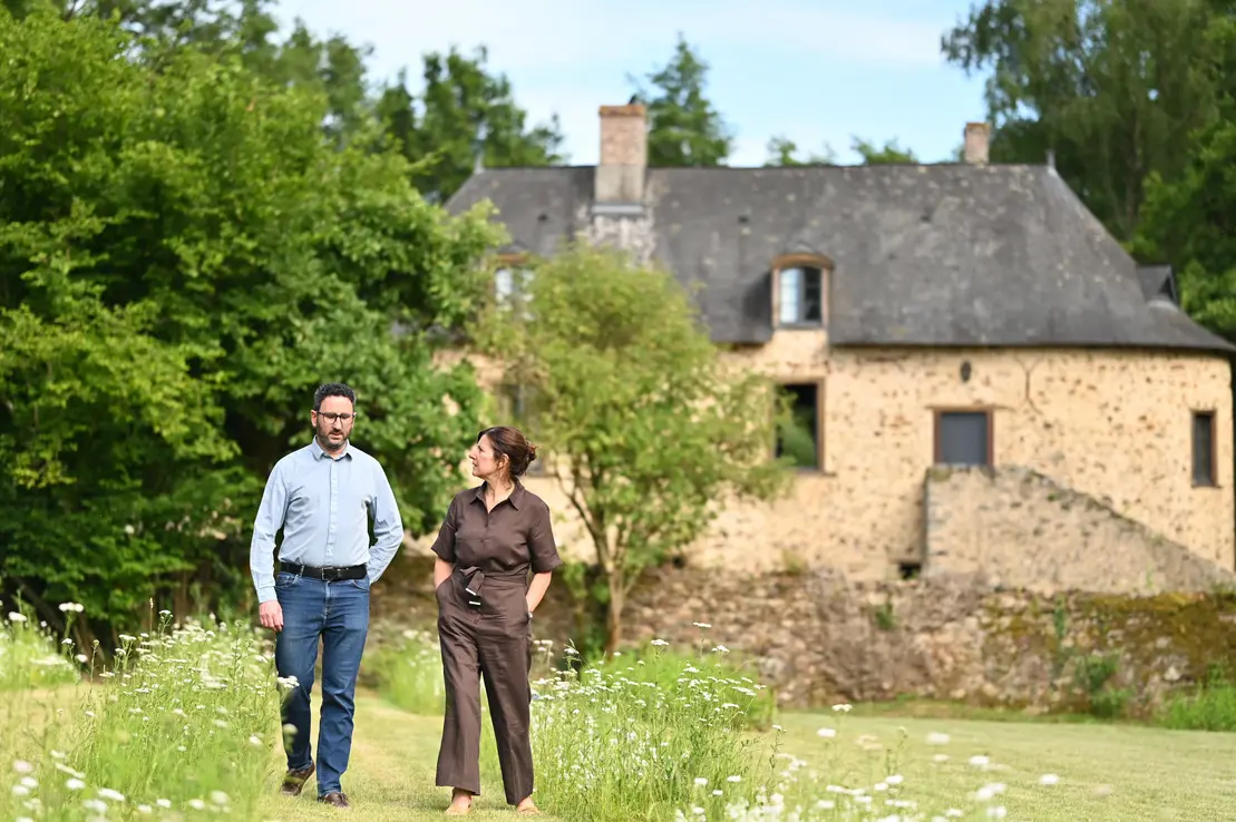 Deux personnes marchant dans une prairie fleurie devant une maison en pierre